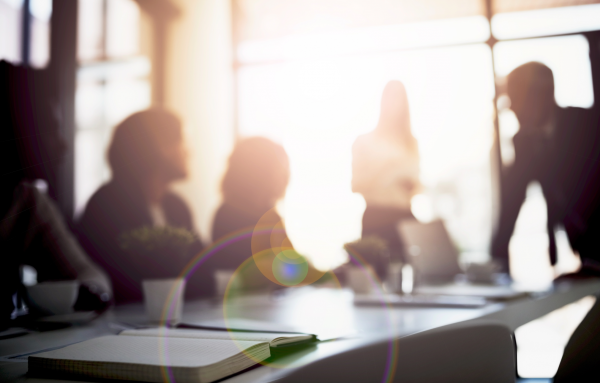 Blurred silhouettes of coworkers meeting around a conference table in a sunlit office with notebooks and laptops visible in the foreground.